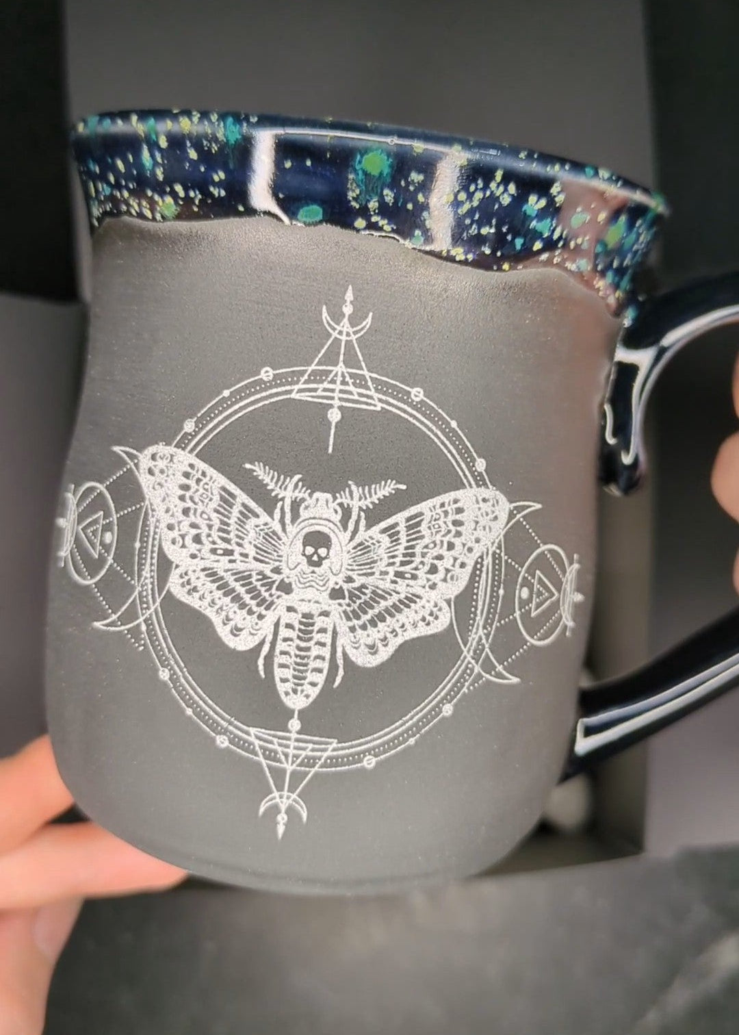 A ceramic mug with a white moth and occult-style line art design on the front, held by hands against a dark background.
